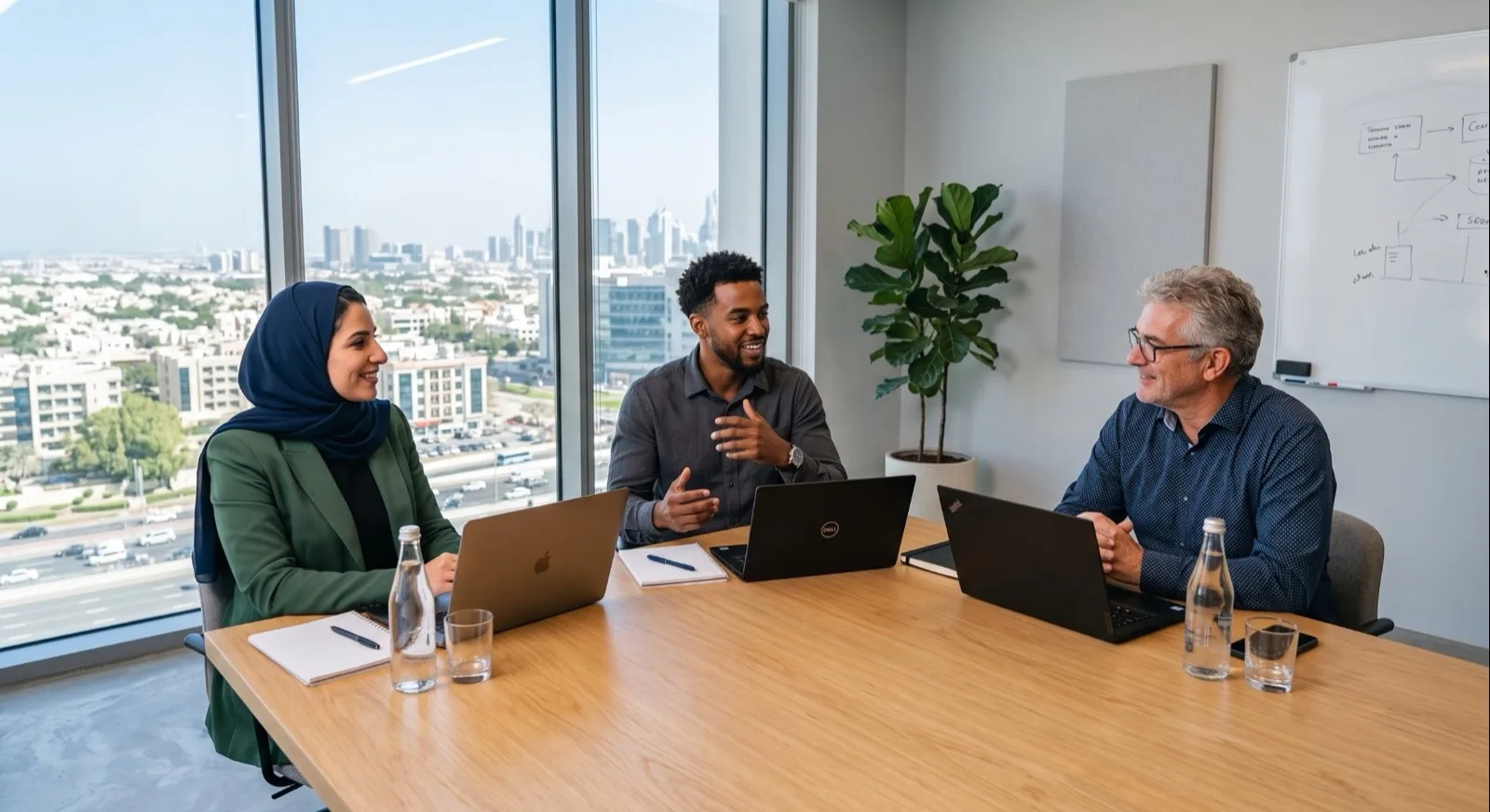Three colleagues talking over laptops at a meeting table in a high-rise office, a city skyline through the window