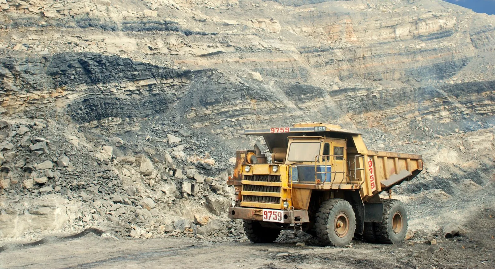 A haul truck parked at the foot of a terraced open-pit mine wall under a cloudy sky