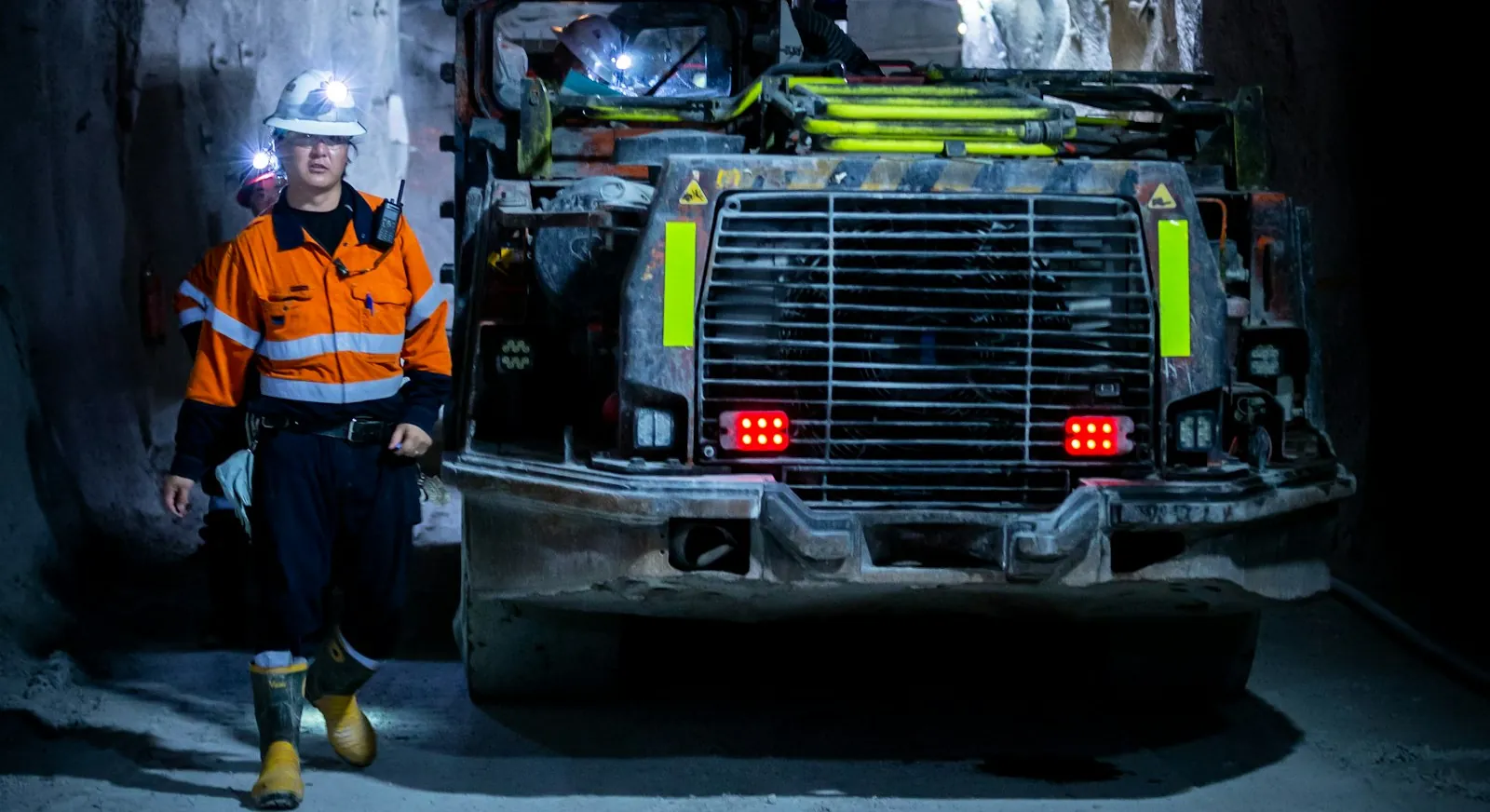 Two underground miners in hard hats with cap lamps walk beside a loader in a mine tunnel