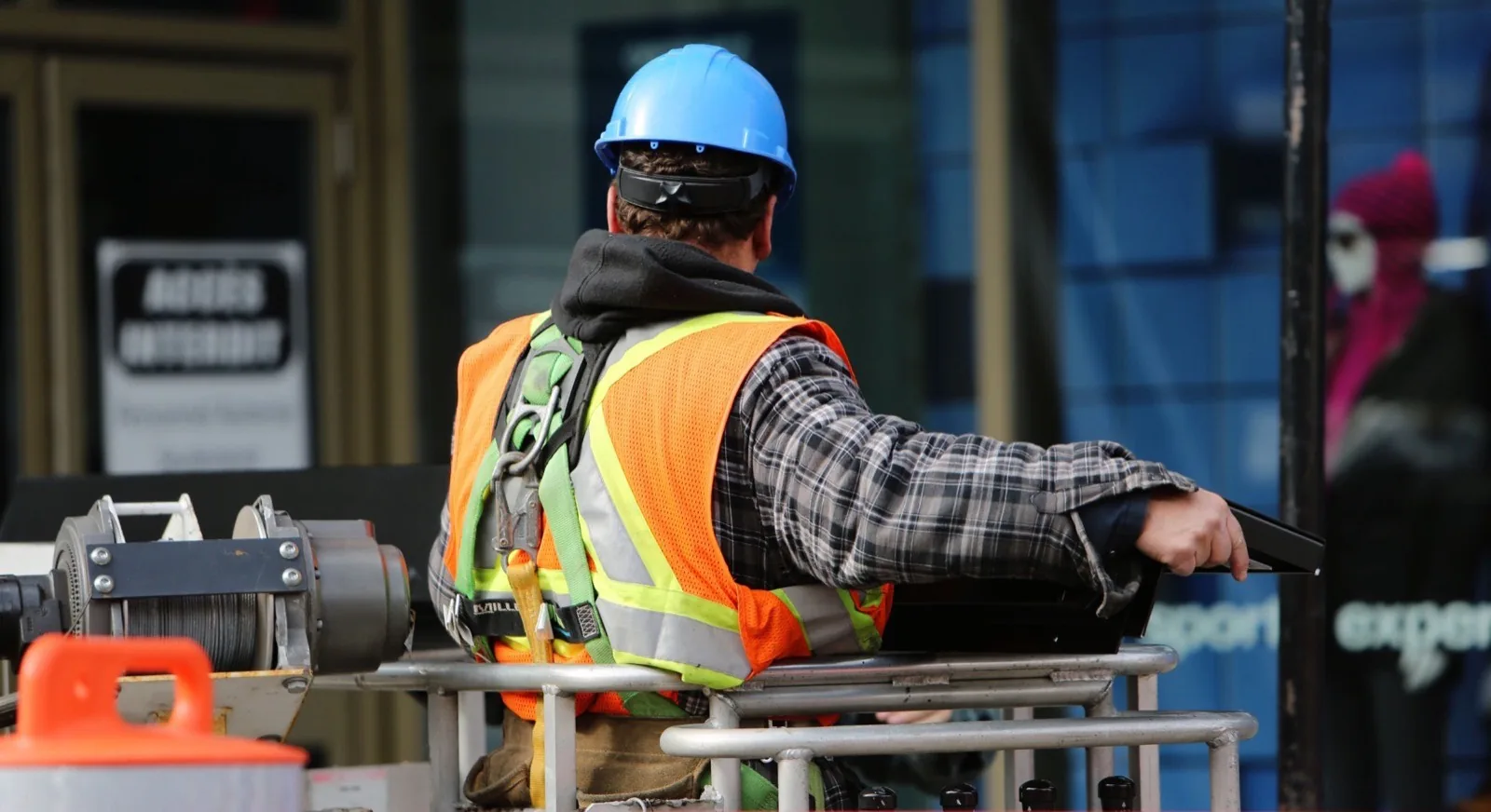 A worker in a hard hat, high-visibility vest and fall-arrest harness at the controls of an elevated work platform