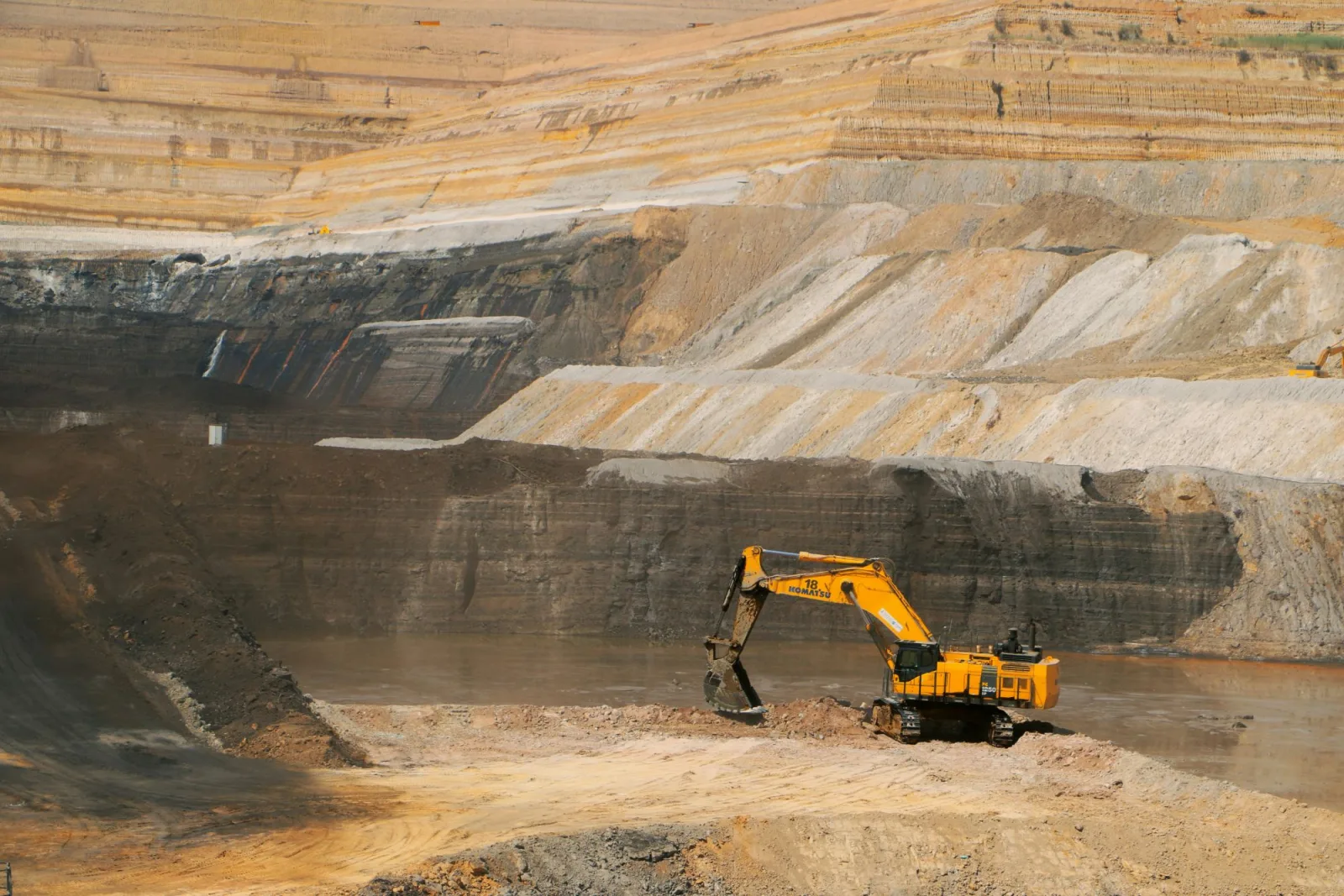 A yellow excavator digs at the floor of a terraced open-pit mine, beside a pool of water