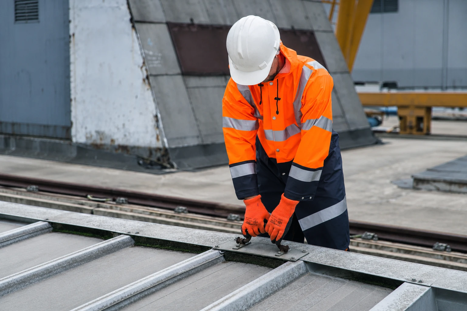 A worker in orange reflective workwear and a white hard hat working on metal fittings at an industrial site